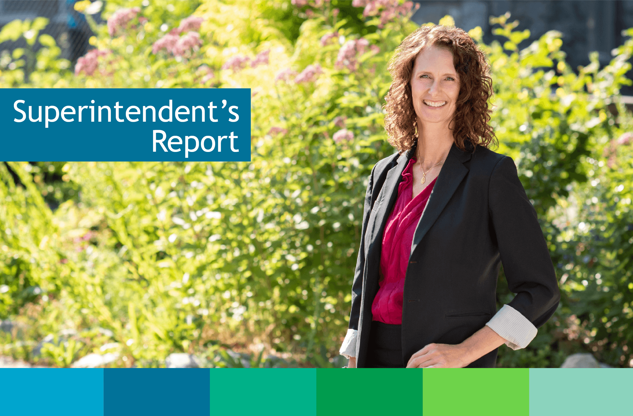 Smiling caucasian woman with brown curly mid-length hair in black blazer and magenta blouse standing in a relaxed pose in front of bright green and flowering bush in the sunshine.