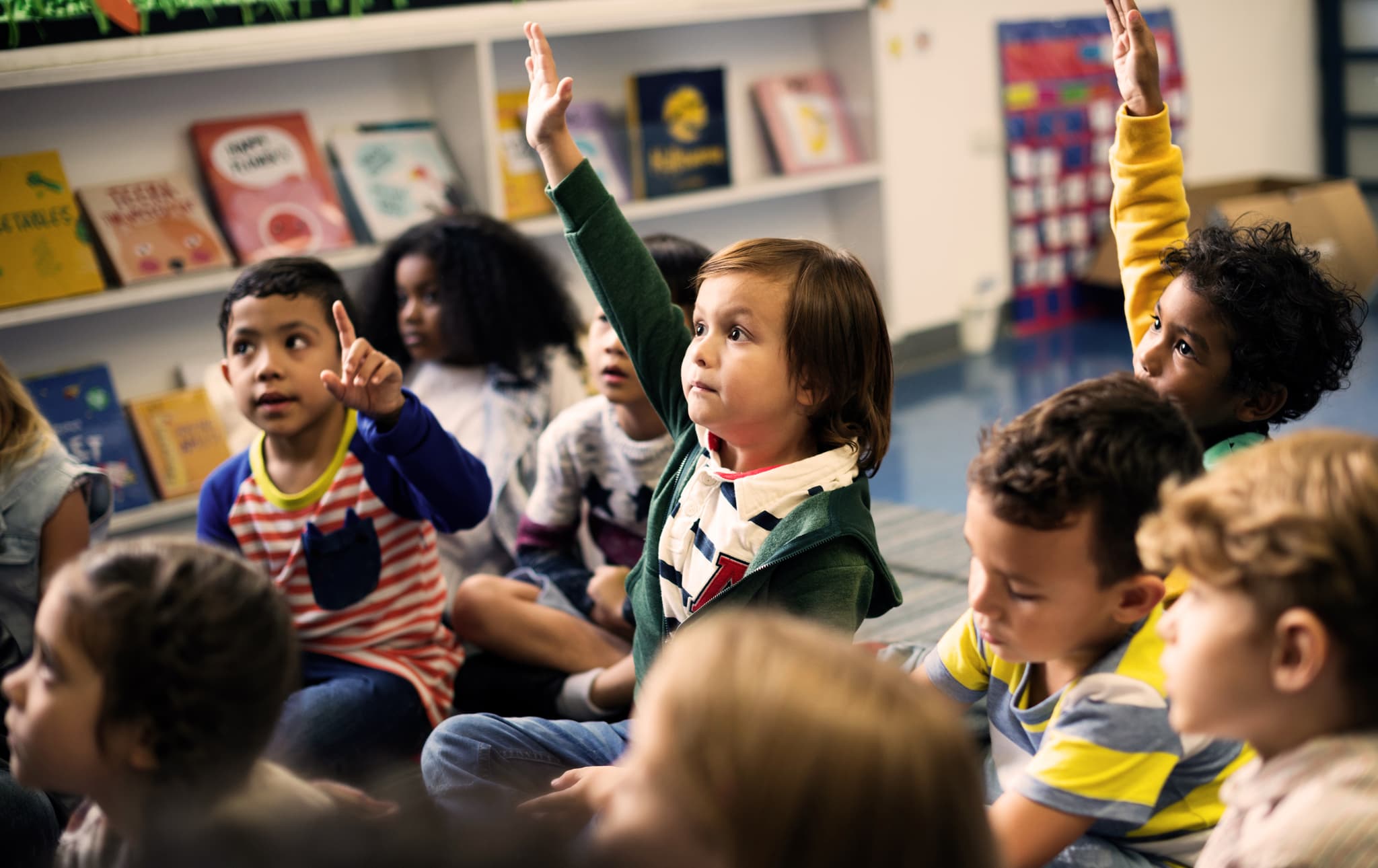 Grade 1 student with hand raised among group of classmates seated on floor.