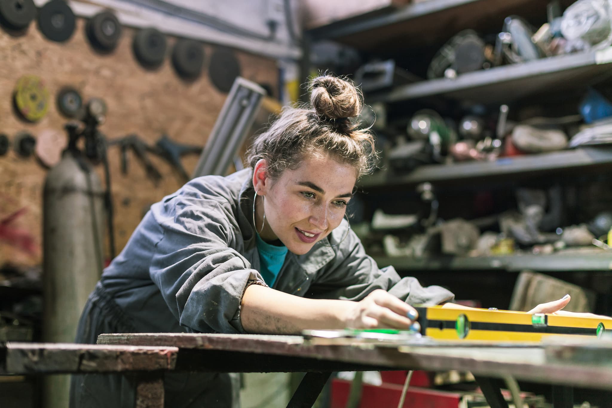 Young woman with hair in top knot wearing coveralls leans over a bench to measure something using a specialized tool