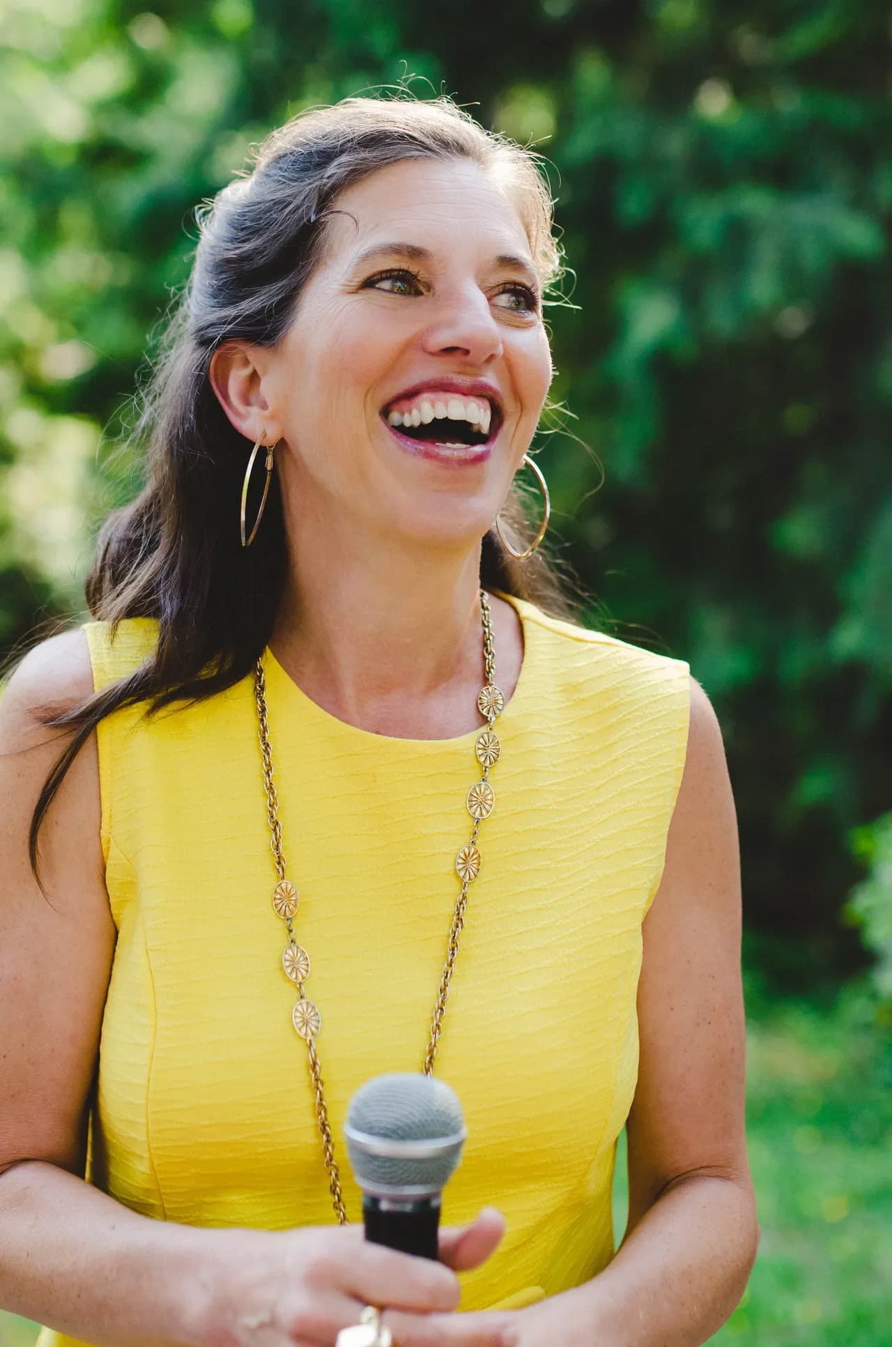 Smiling woman with long brown hair in yellow sleeveless dress holds a microphone while standing in front of green bushes and green grass
