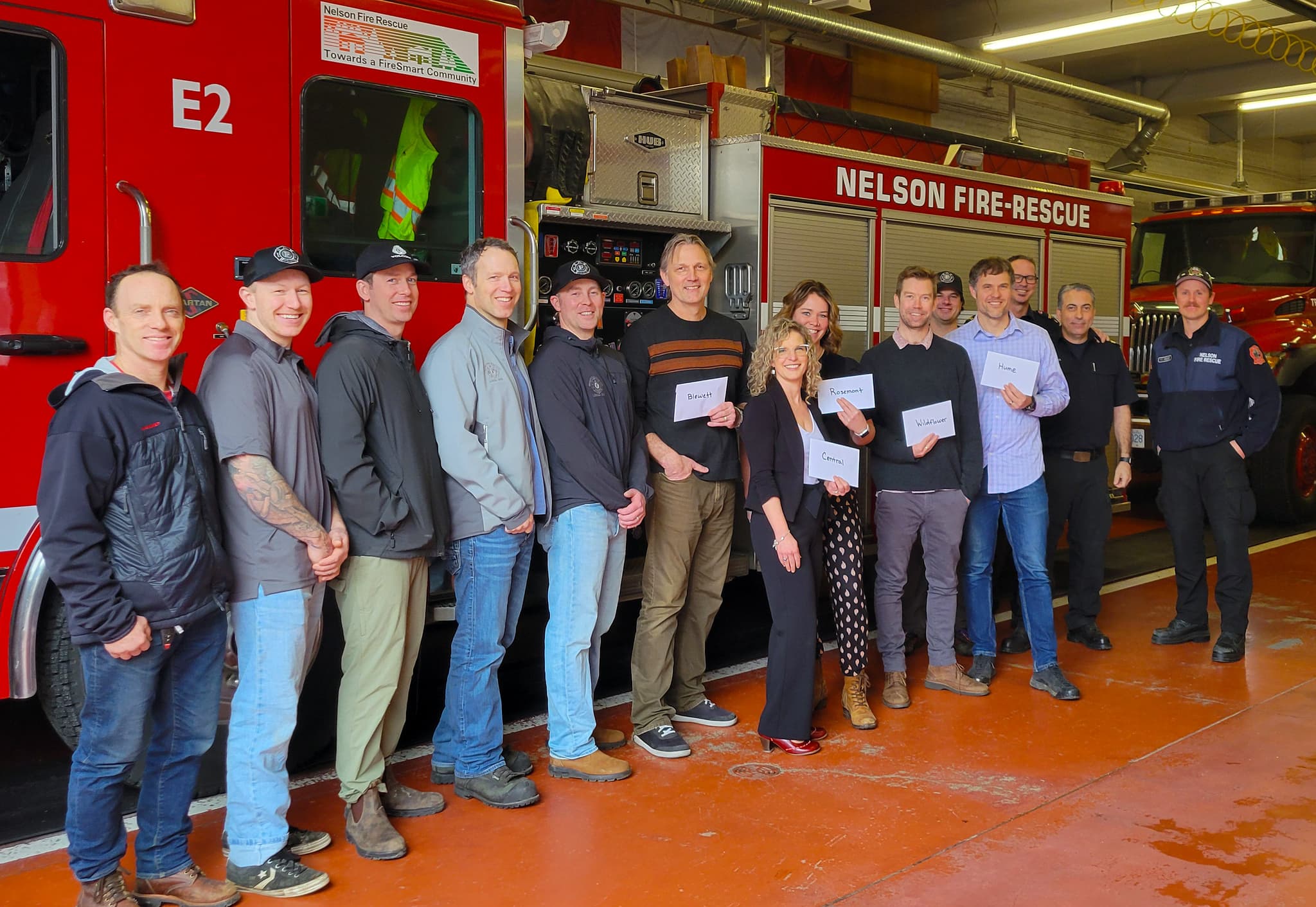 A group of people lined up in front of a fire truck inside the bay of a fire department smiling, with some holding envelopes and some in fire fighter uniforms.