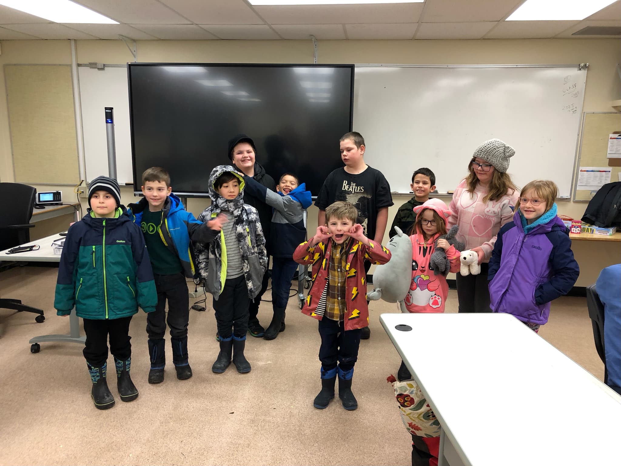 Elementary school children standing in a classroom with winter jackets, boots and toques on, making faces, smiling, watching.