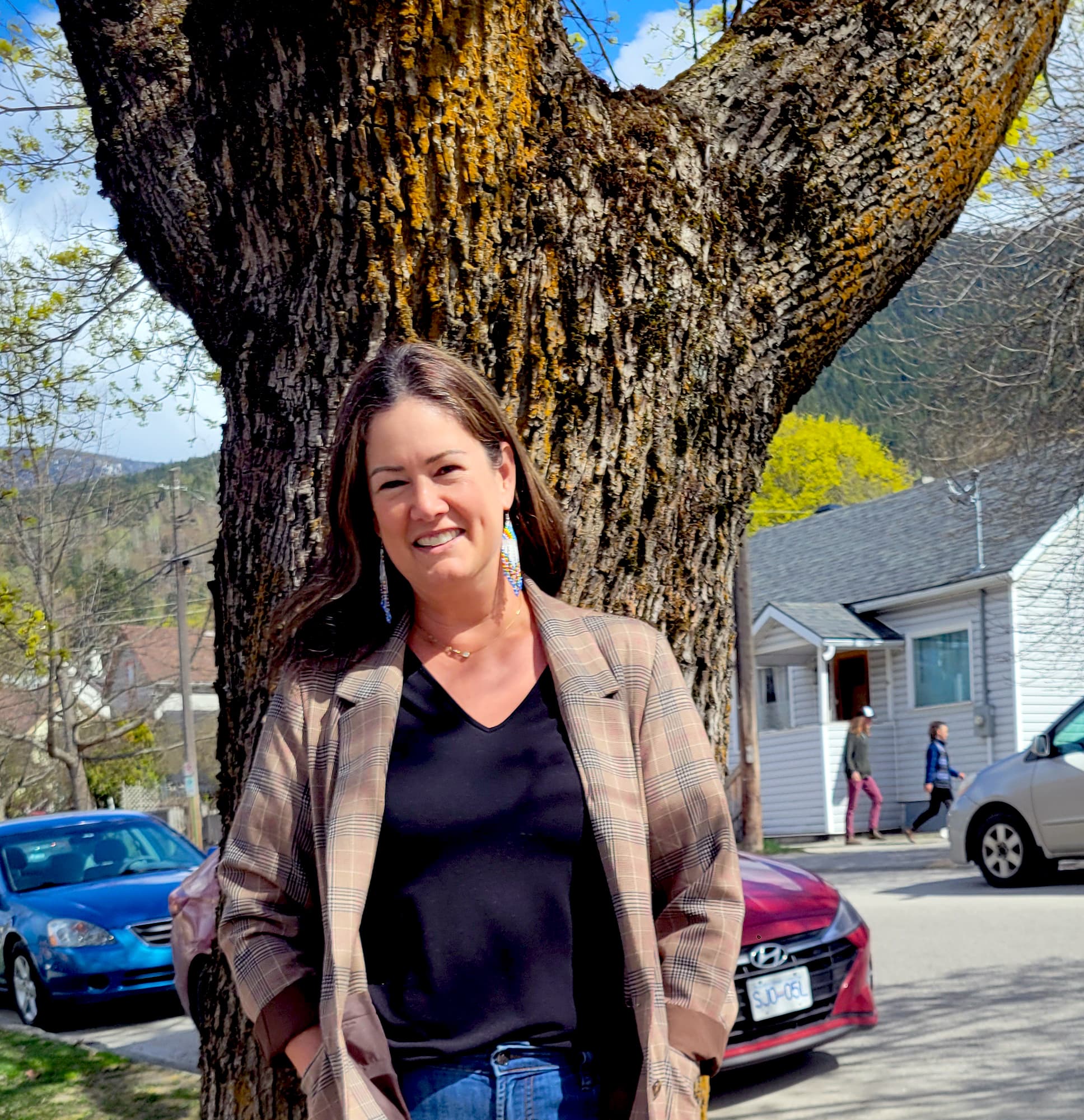 Woman with brown hair in tweed blazer, black shirt and blue jeans standing in front of the trunk of a maple tree on a sunny day.