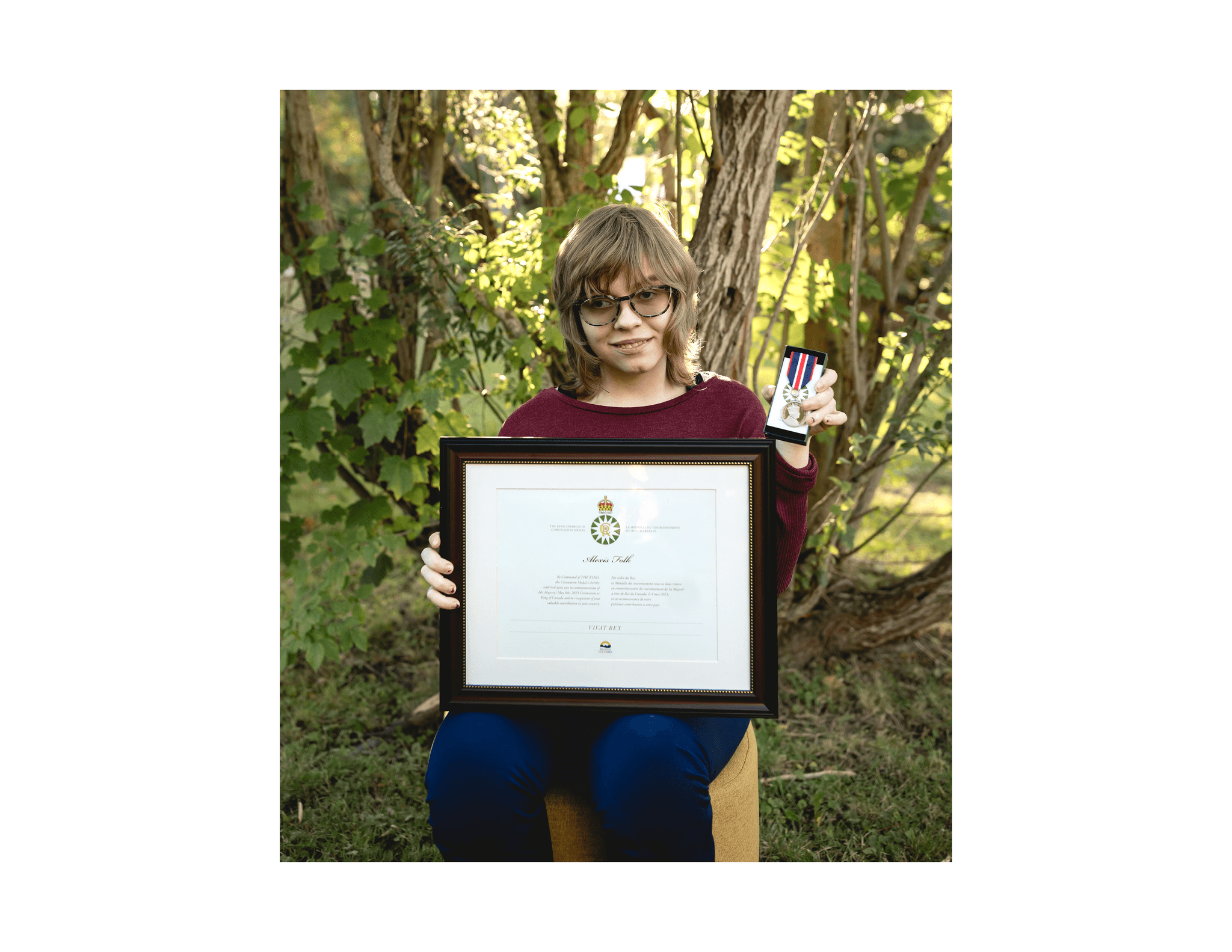 A young woman with wavy blonde hair wearing a burgundy sweater seated on a chair in a forest holding a medal and a framed certificate.