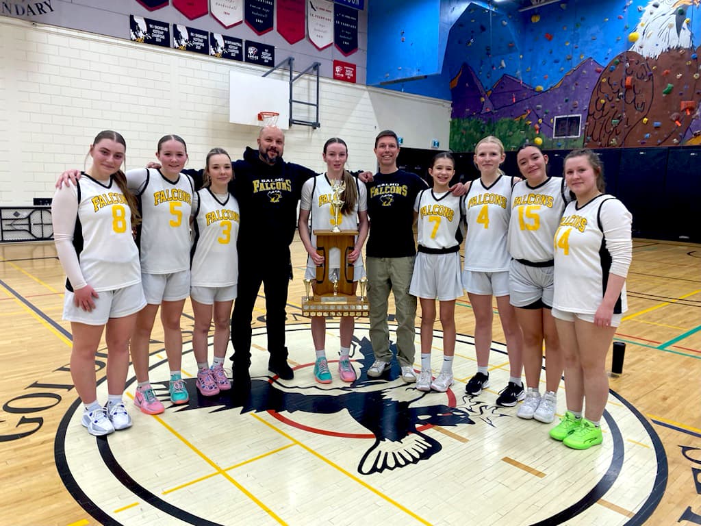 A highschool girls' basketball team stands with their coaches, holding a large trophy in the centre of a gymnasium.
