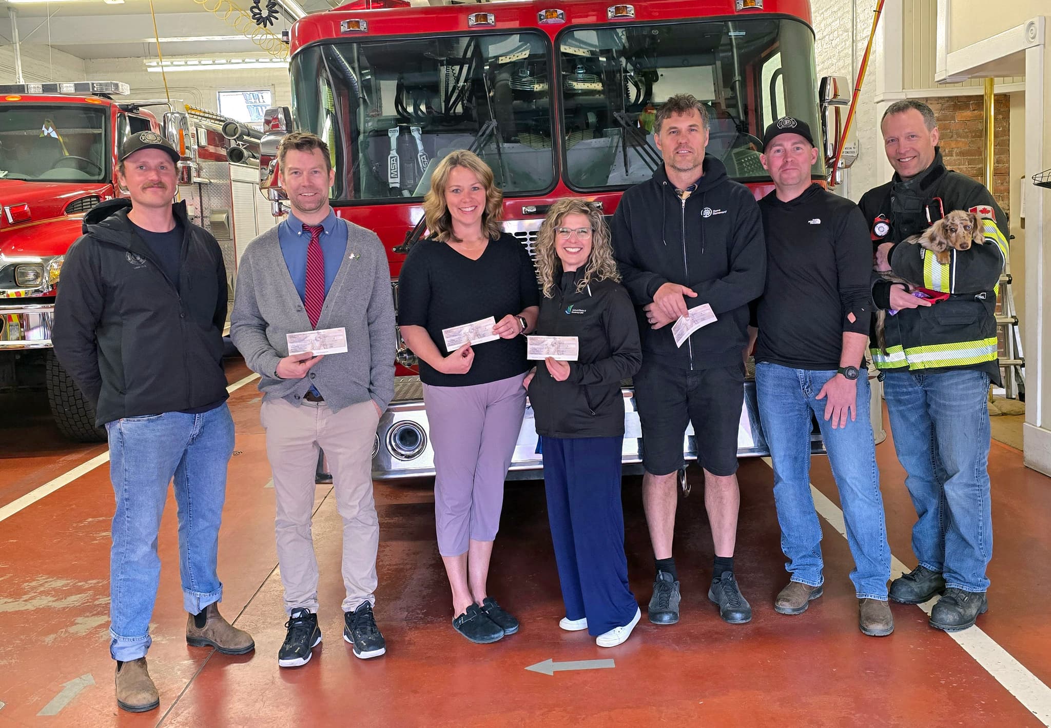 A group of people stand in front of a fire truck inside a fire department, holding cheques in front of themselves.