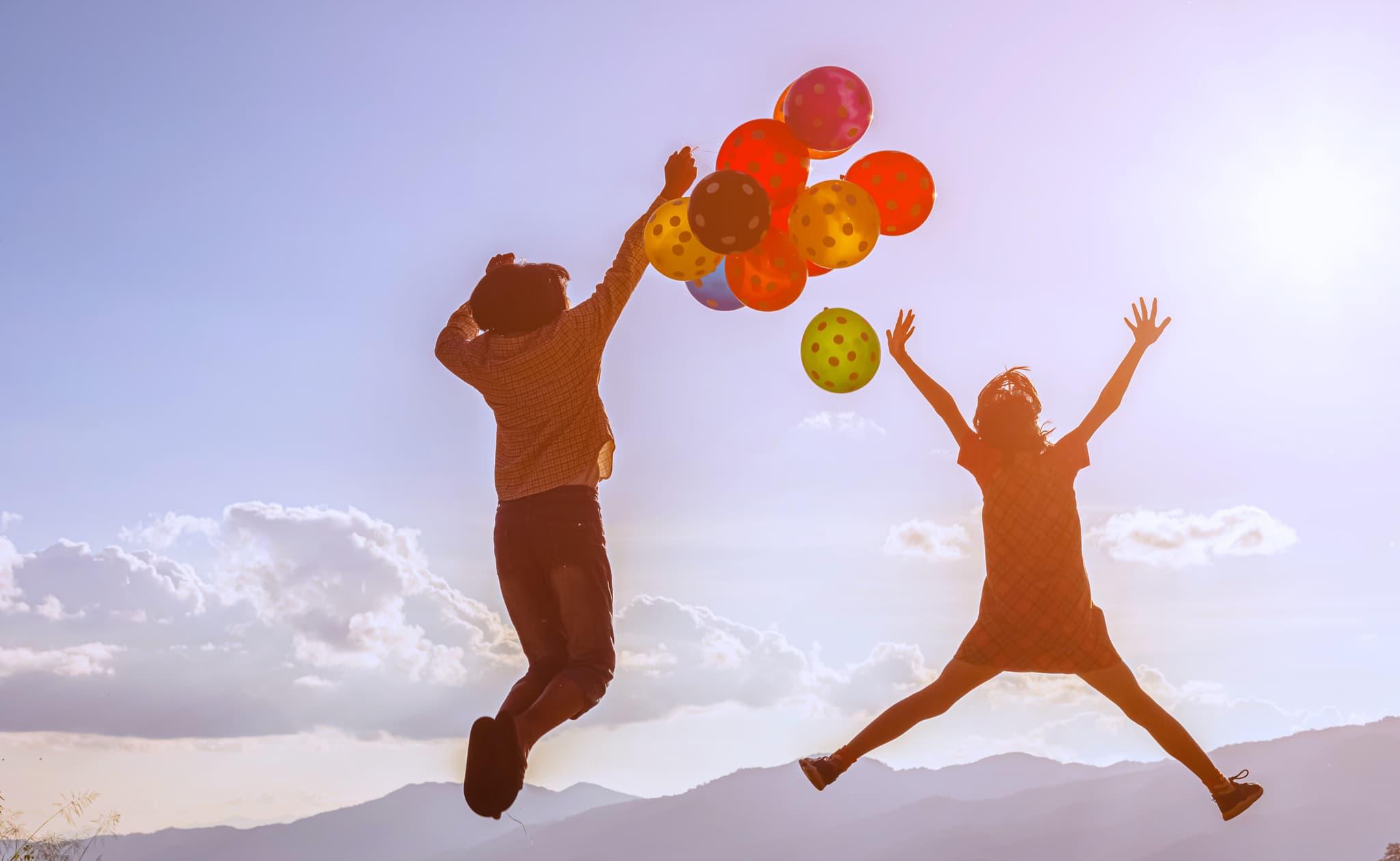 Two children leap into the air with coloured balloons against a background of mountains and sunshine.