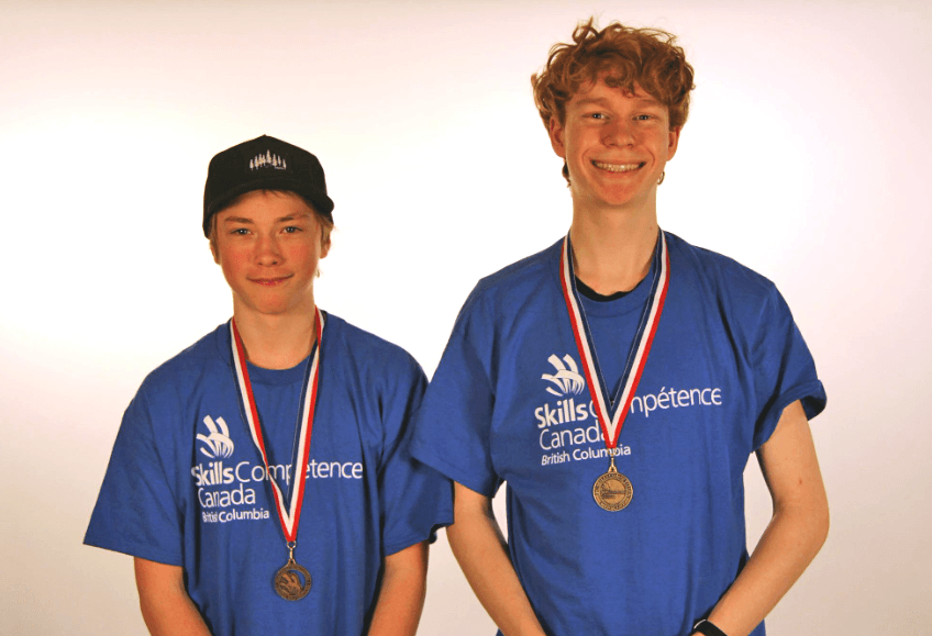Two teenaged boys in blue t-shirts wearing gold medals and standing in front of a white background.