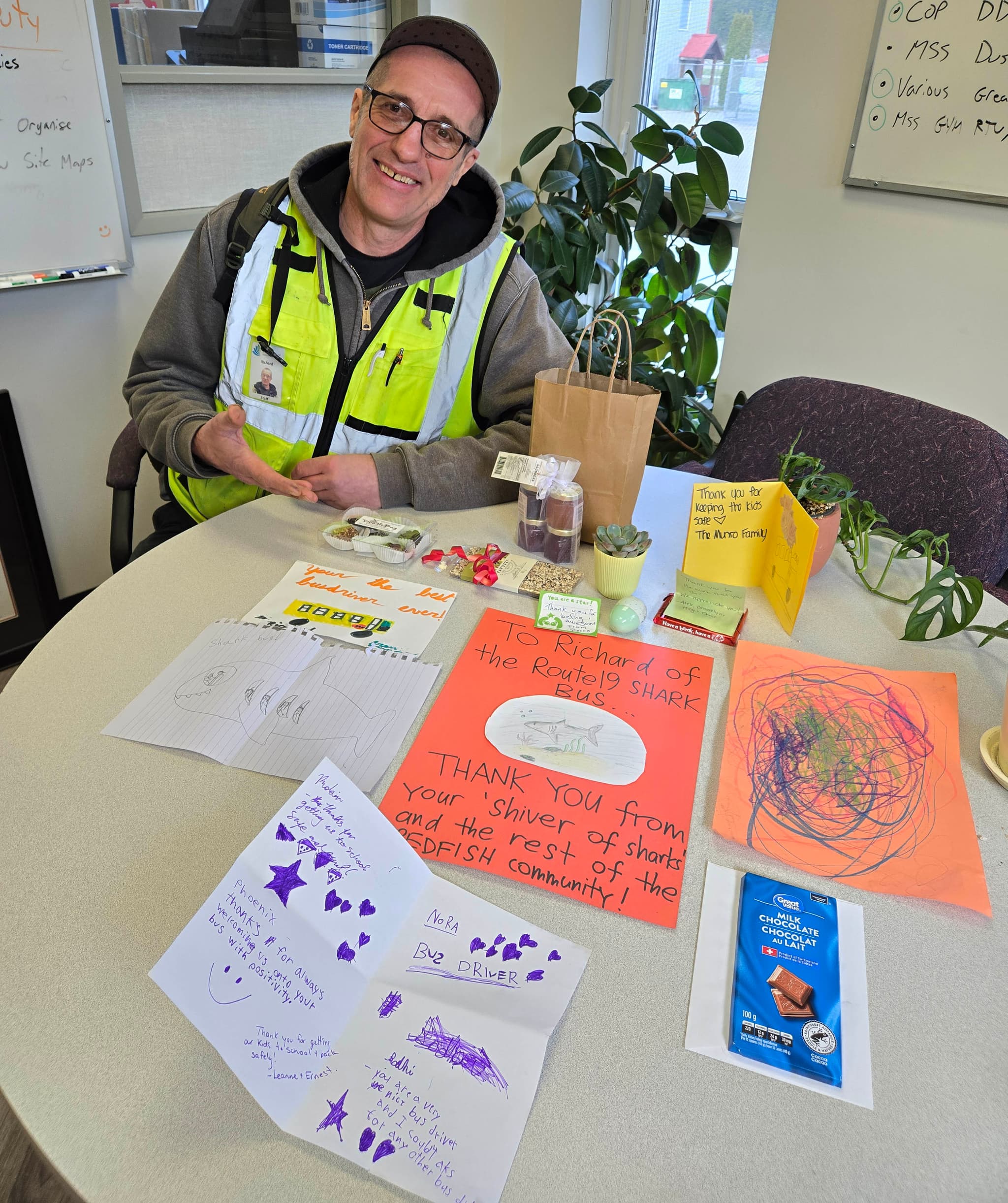 A man in a ball cap, glasses and a flourescent green vest sits at a table covered in hand-drawn cards and messages.
