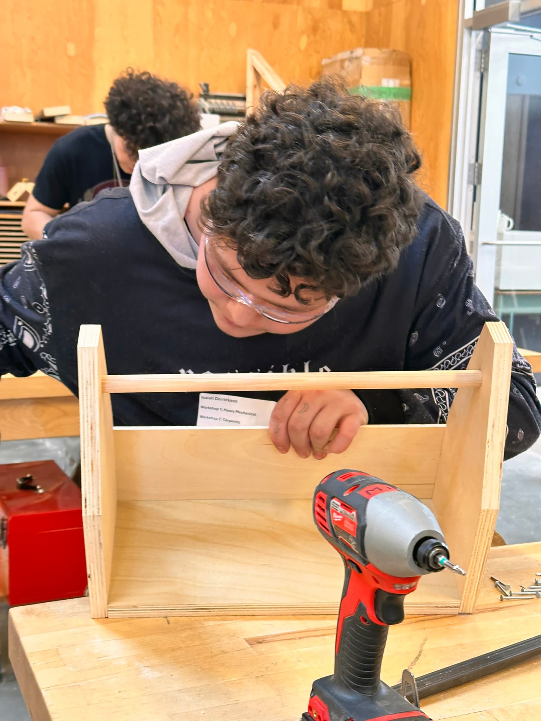 A teenager leans over a workbench working on a woodworking project with a red drill in the foreground and another teenager working on a project behind him.