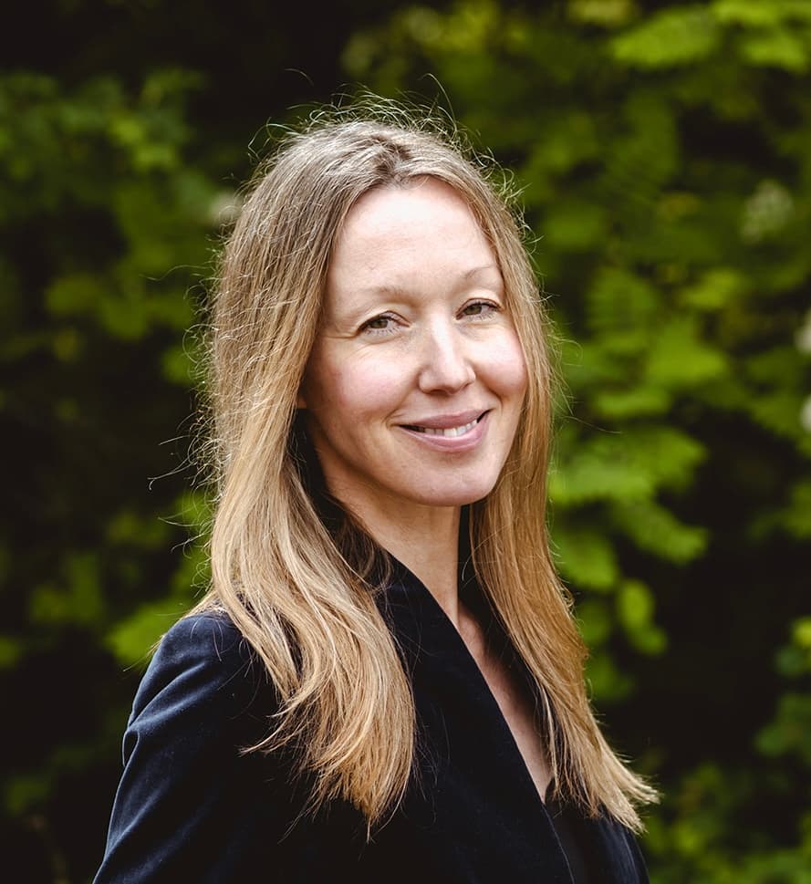 A woman with dark blonde long hair wearing a black shirt smiles in front of a green bush outdoors.