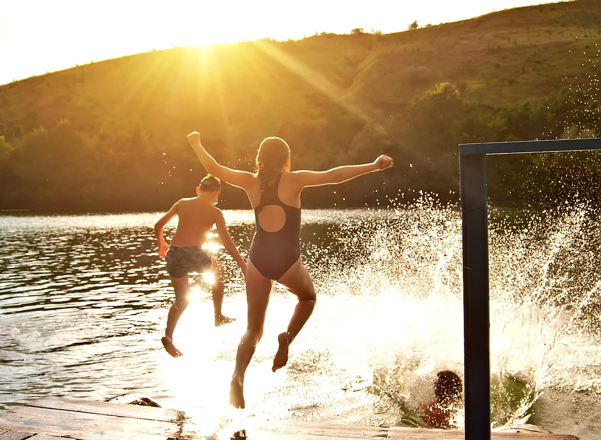 Three children leap off a dock into a lake at sunset