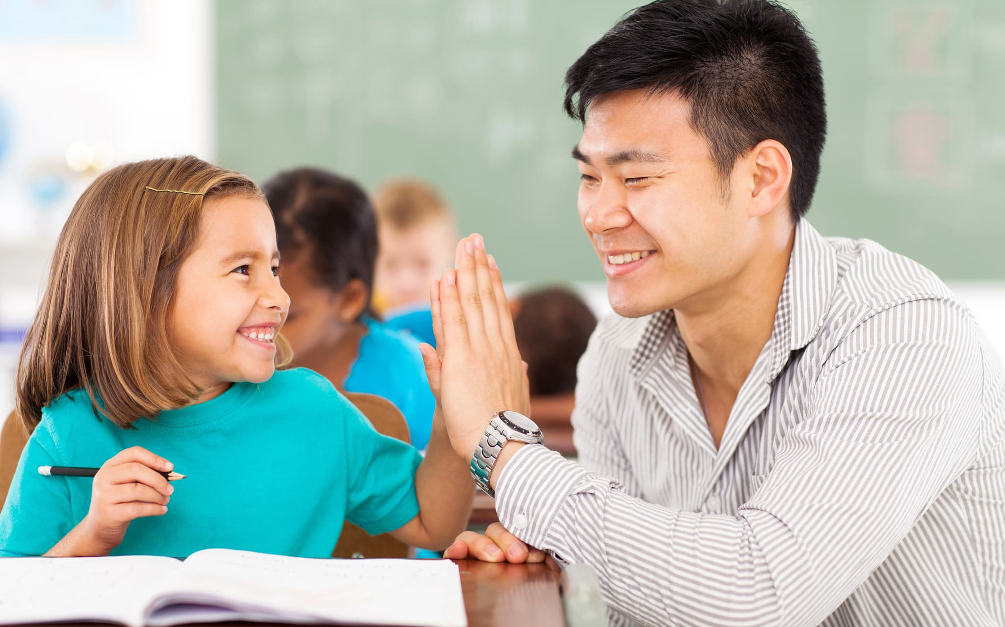 A young elementary student sitting at her desk high fives her male teacher who is crouched beside her. Both are smiling widely.