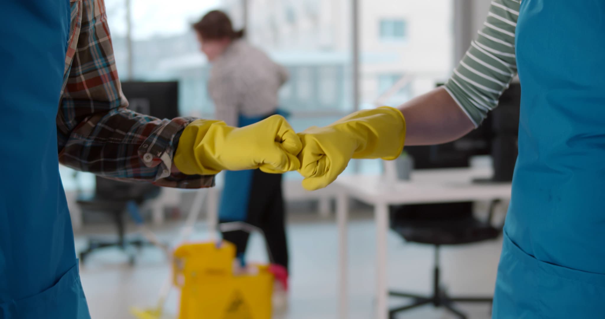 Custodians wearing yellow rubber gloves and blue aprons bump fists in an office setting while a custodian mops in the background