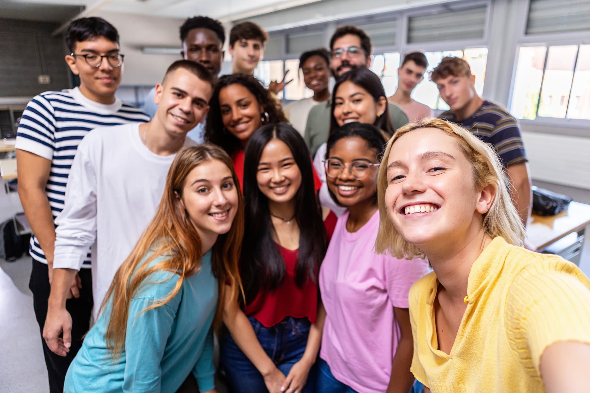Selfie taken of smiling, diverse group of teens in classroom with teacher in background