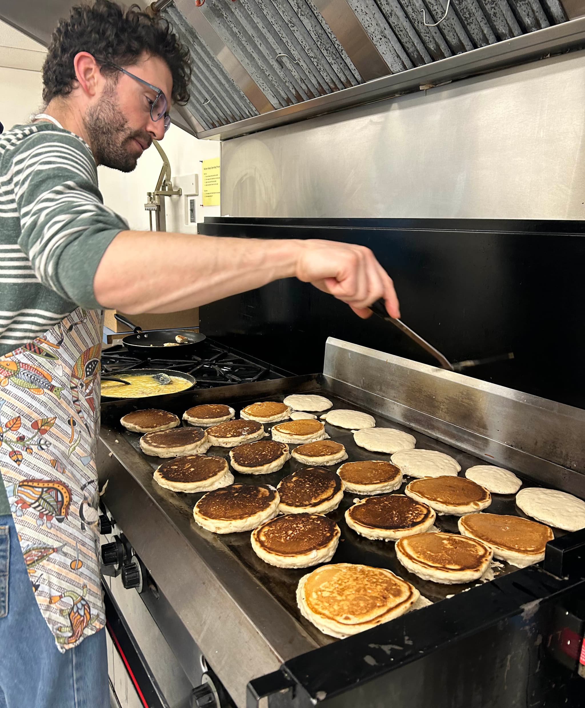 A man with brown hair, a close beard and glasses wearing a colourful apron cooks pancakes on a commercial grill.