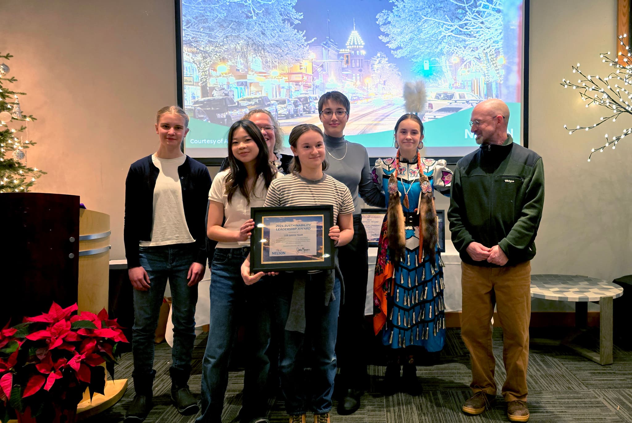 A group of young people holding a framed award posing in front of a screen and flanked to the right by an adult looking on.