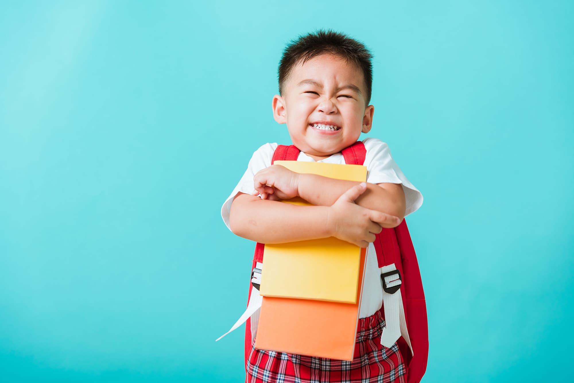 A preschool child with short black, spiky hair wearing a backpack and hugging some paper school supplies while he smiles in excitement.