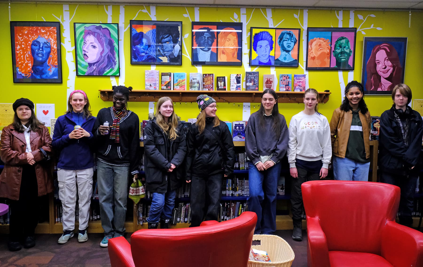 High school students stand in front of a yellow wall displaying their colourful artwork, with two red, leather chairs in the foreground.
