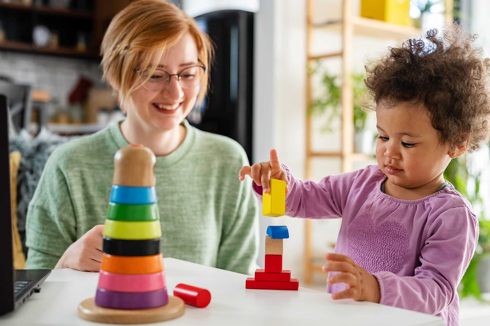 Young woman looks on, smiling, as a toddler builds a tower of coloured blocks.