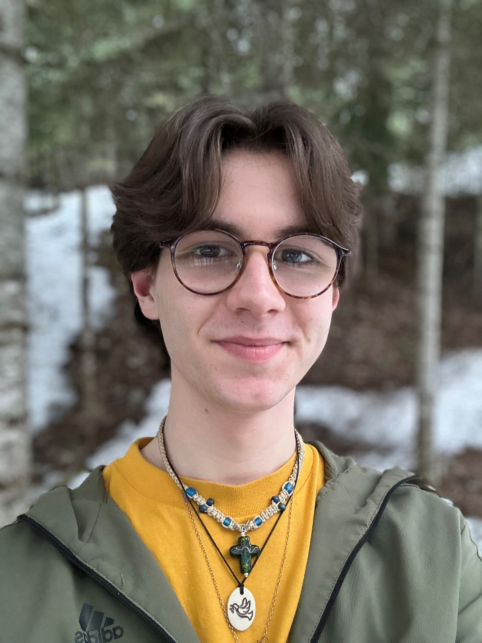 A teenaged boy with brown hair wearing glasses and a green jacket stands in front of some trees.