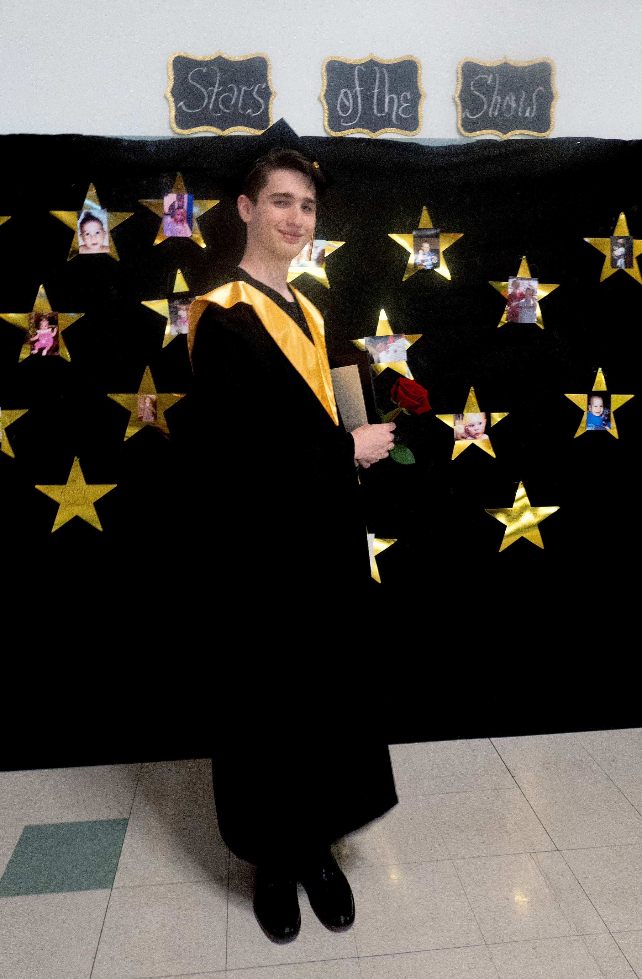A high school graduate in cap and gown standing in front of a black backdrop decorated with gold stars.