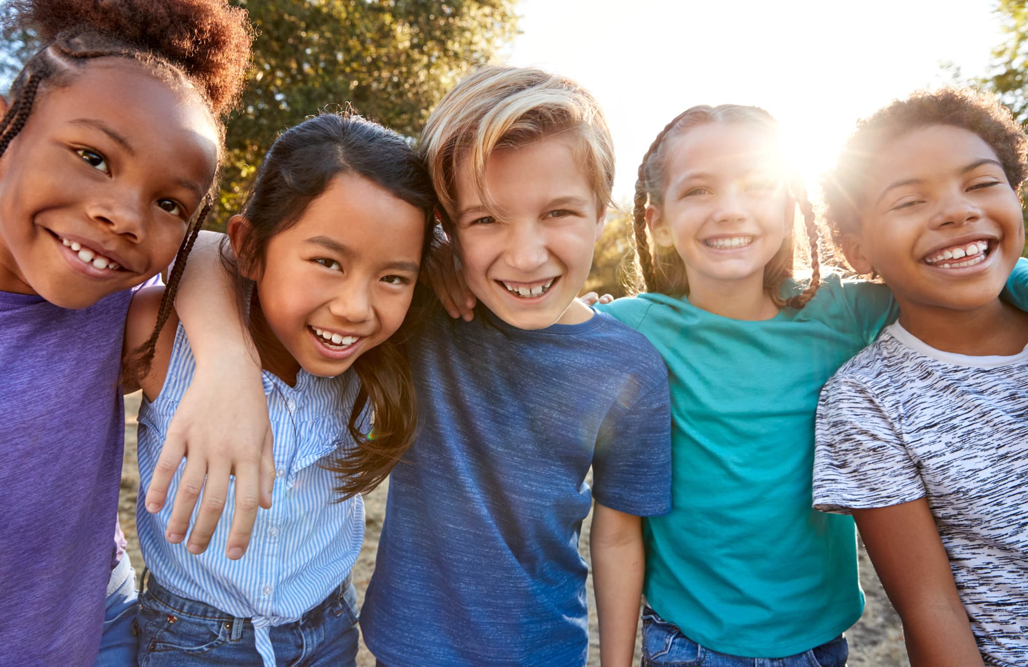 A group of diverse children arm in arm against a backdrop of sunlit trees