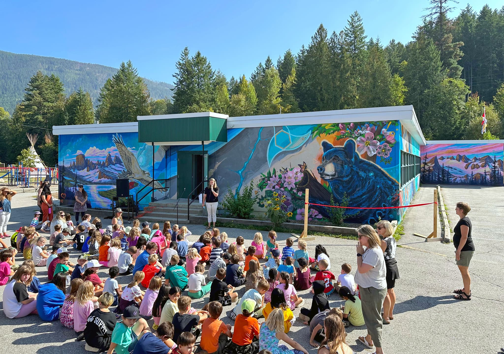 A group of elementary school students sit on the ground in front of a colourful school while a principal speaks to them.