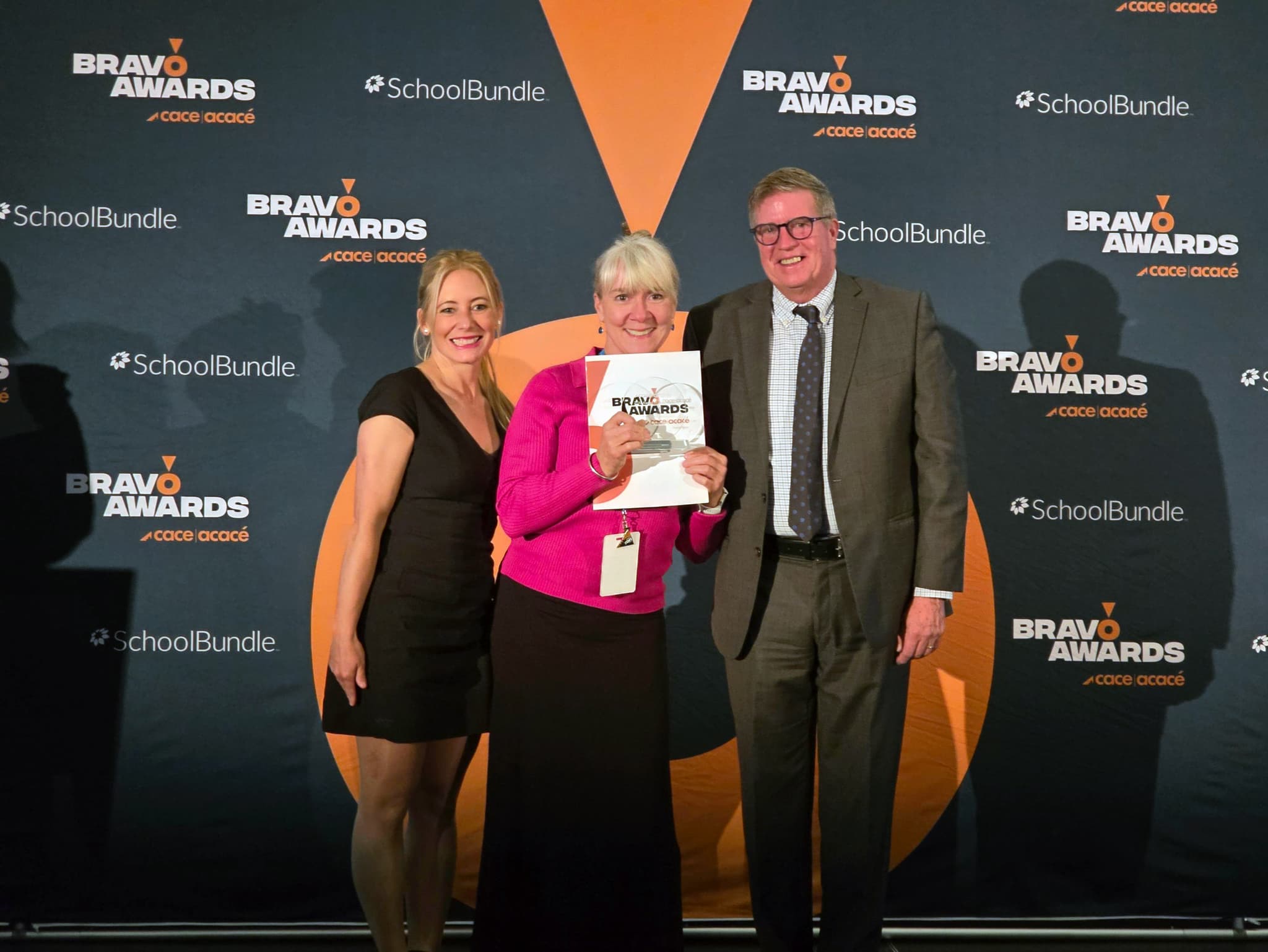 Two women in dresses and a man in a suit pose in front of a grey wall with the middle woman holding up a certificate.