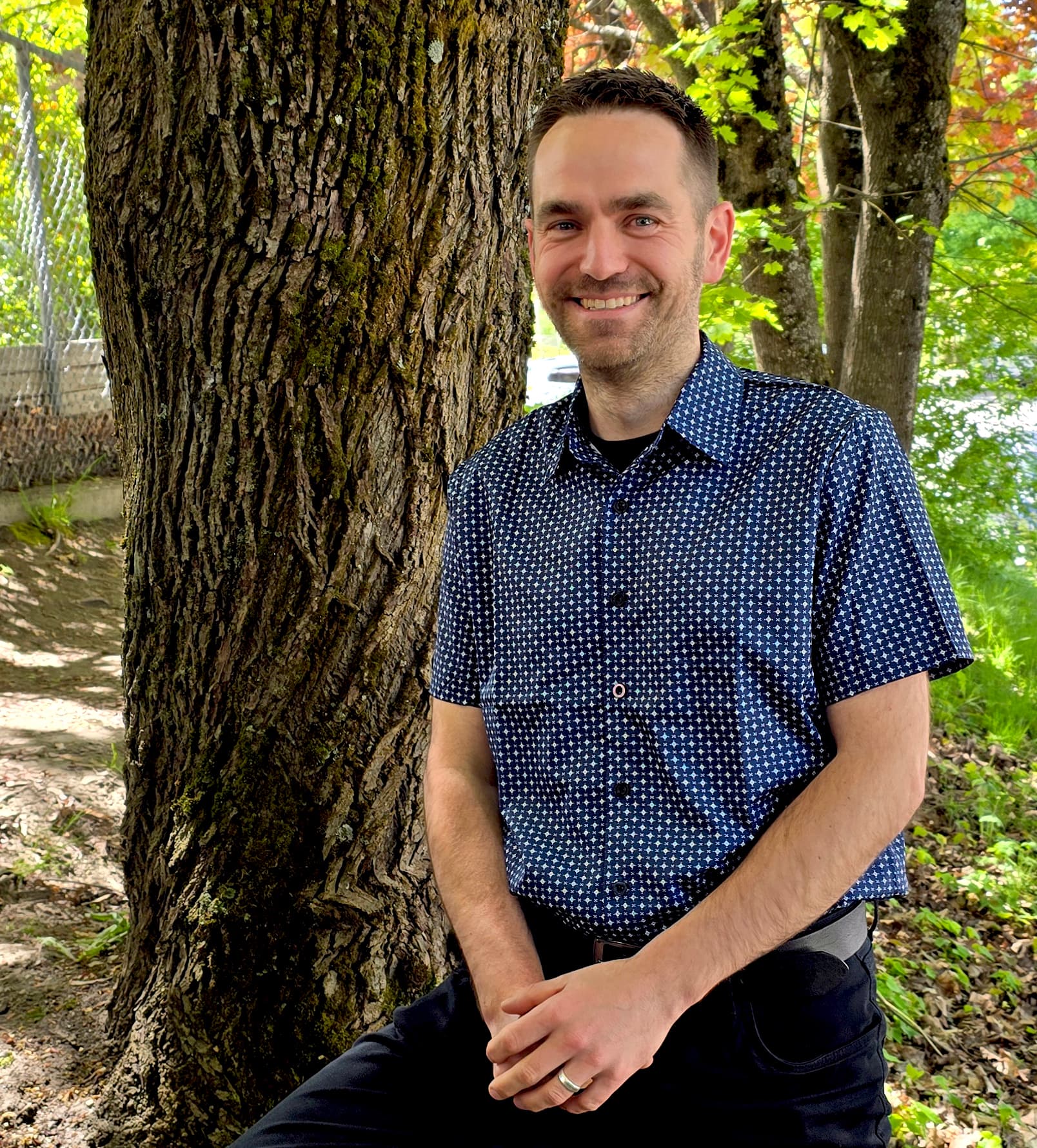 A man with short blonde-brown hair in a blue short-sleeved button-up shirt stands smiling in front of a tree in the sunshine.