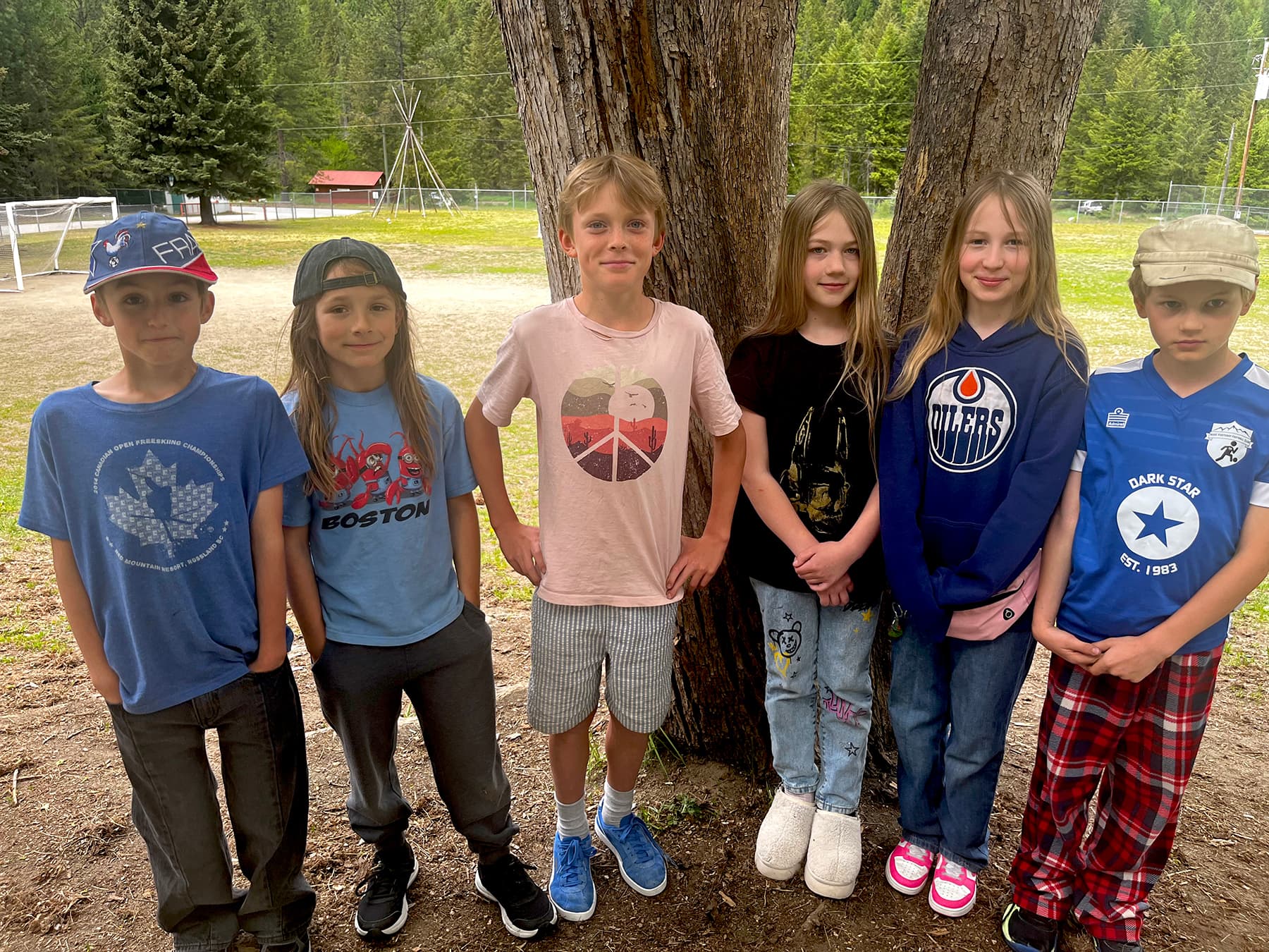 Six children stand outside in front of two trees with a school playground behind them.