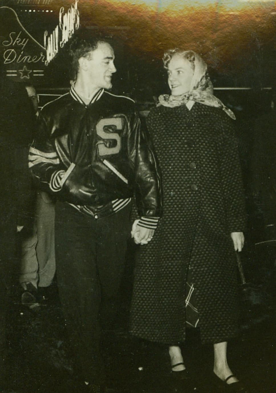 A black and white photo from the 1950s of a man in a letter jacket and a women in a long coat and kerchief smiling at one another and walking at night while holding hands