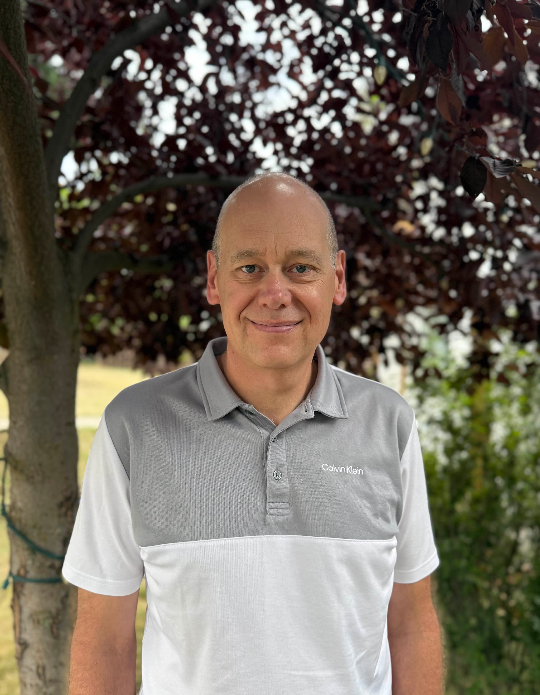 A man who is mostly bald wearing a grey and white short sleeved golf shirt standing and smiling in front of a leafy tree