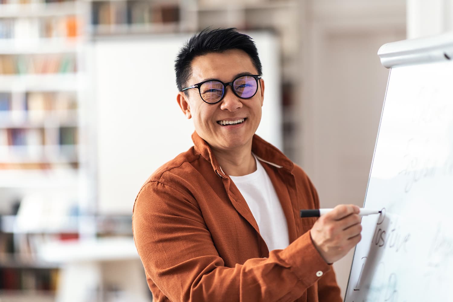 Man with black hair and black glasses teacher smiling writing on whiteboard in classroom